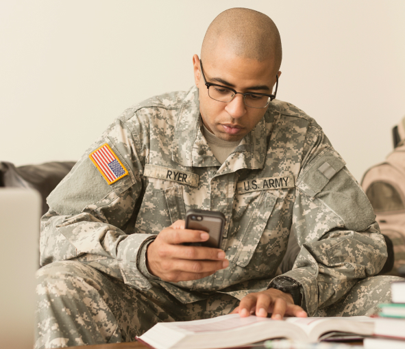 A man in fatigues looks at his phone while studying a book