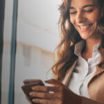 Woman smiling at her smartphone near a large window.