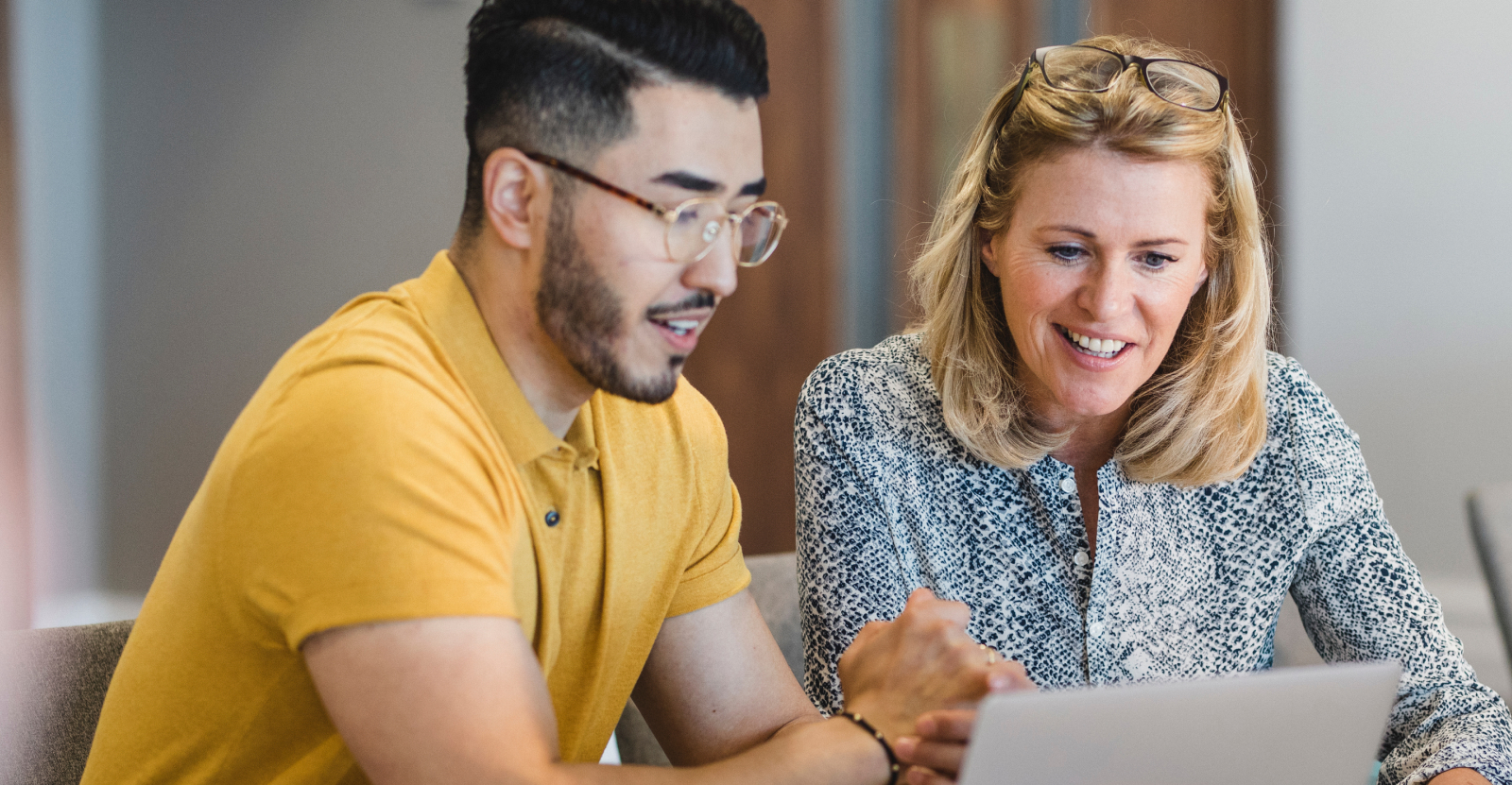 Two people looking at a laptop together, one wearing a yellow shirt and glasses, the other a blue-patterned blouse.