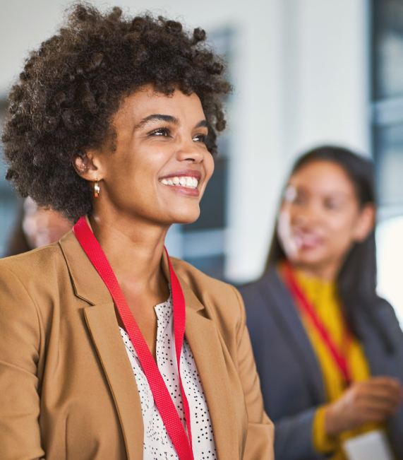 A woman with curly hair smiles, wearing a tan blazer and red lanyard, in a bright room.