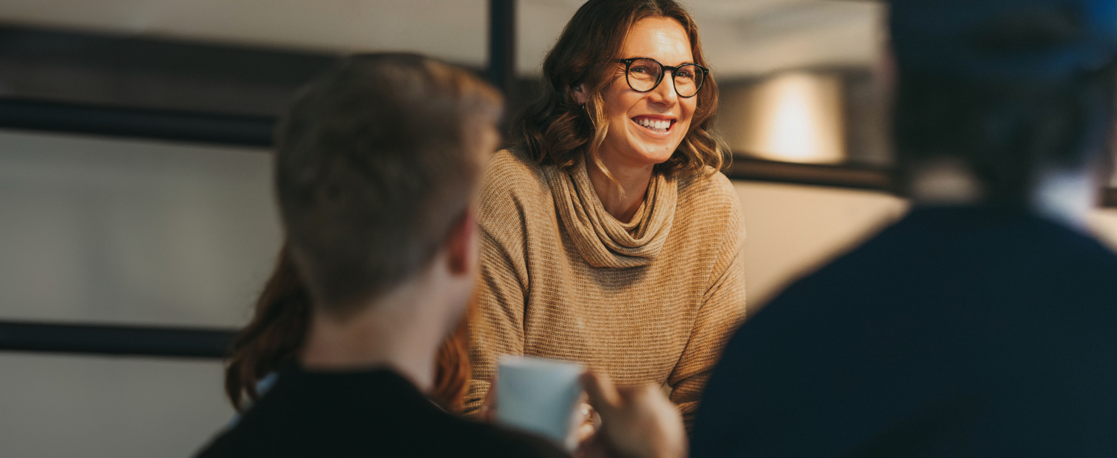 A smiling person in glasses and a light brown sweater, engaged in conversation, with two others in the foreground.