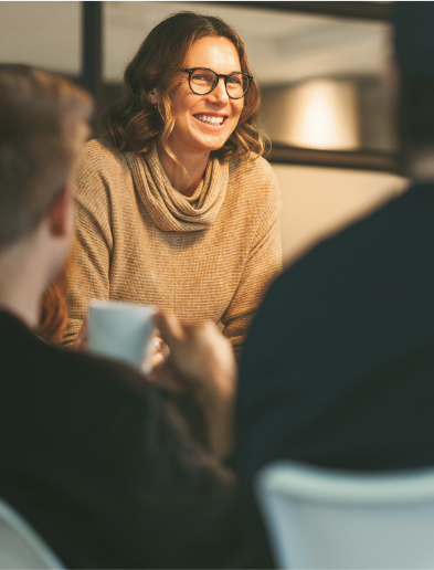 A smiling person in glasses and a light brown sweater, engaged in conversation, with two others in the foreground.