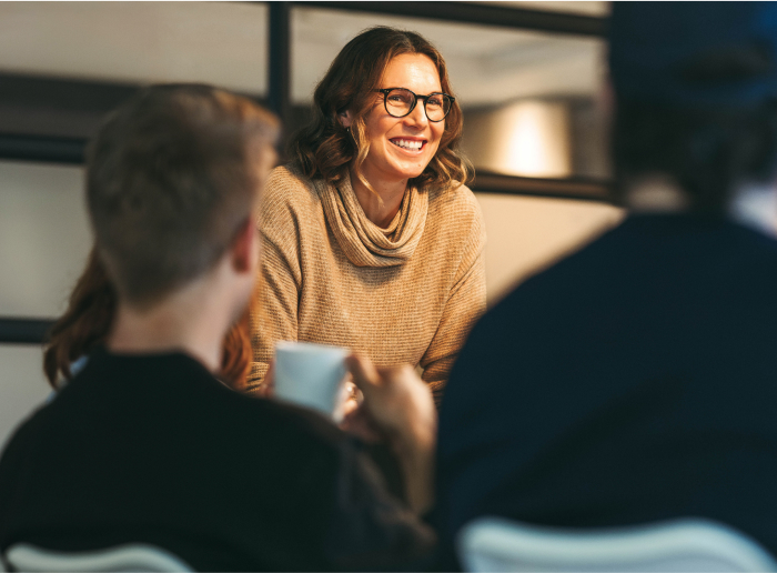 A smiling person in glasses and a light brown sweater, engaged in conversation, with two others in the foreground.