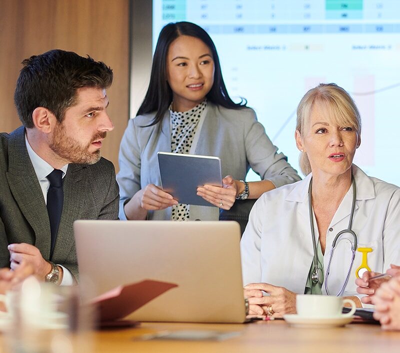 A group of healthcare professionals and a consultant at a table with a spreadsheet on a monitor on the wall.