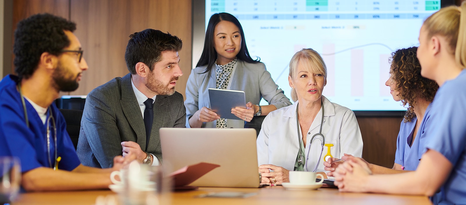 A group of healthcare professionals and a consultant at a table with a spreadsheet on a monitor on the wall.