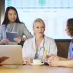 A group of healthcare professionals and a consultant at a table with a spreadsheet on a monitor on the wall.