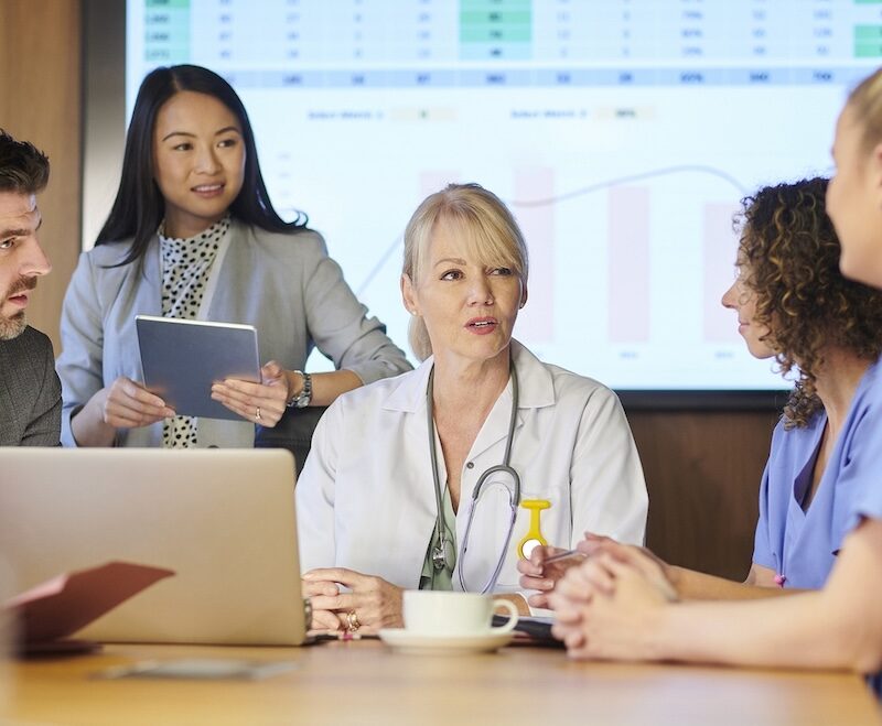 A group of healthcare professionals and a consultant at a table with a spreadsheet on a monitor on the wall.