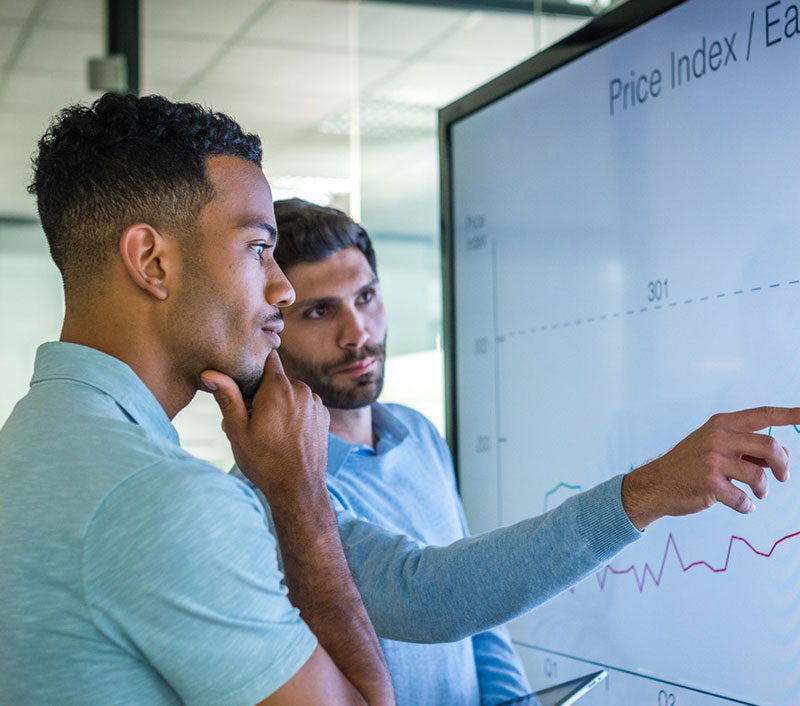 Two men standing in front of a large monitor looking at a upward trending line graph.