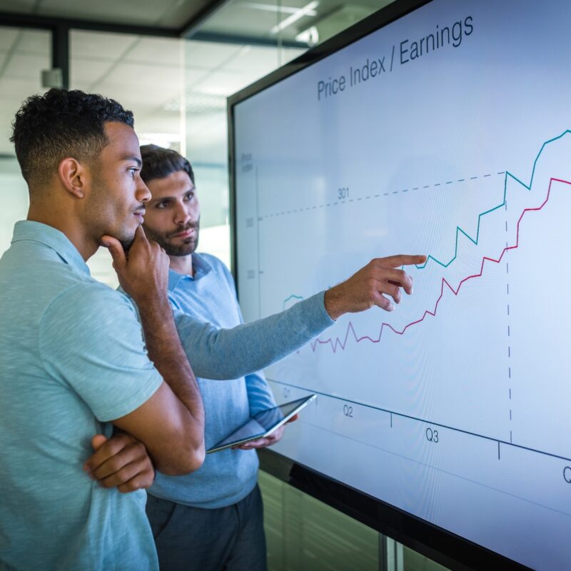 Two men standing in front of a large monitor looking at a upward trending line graph.