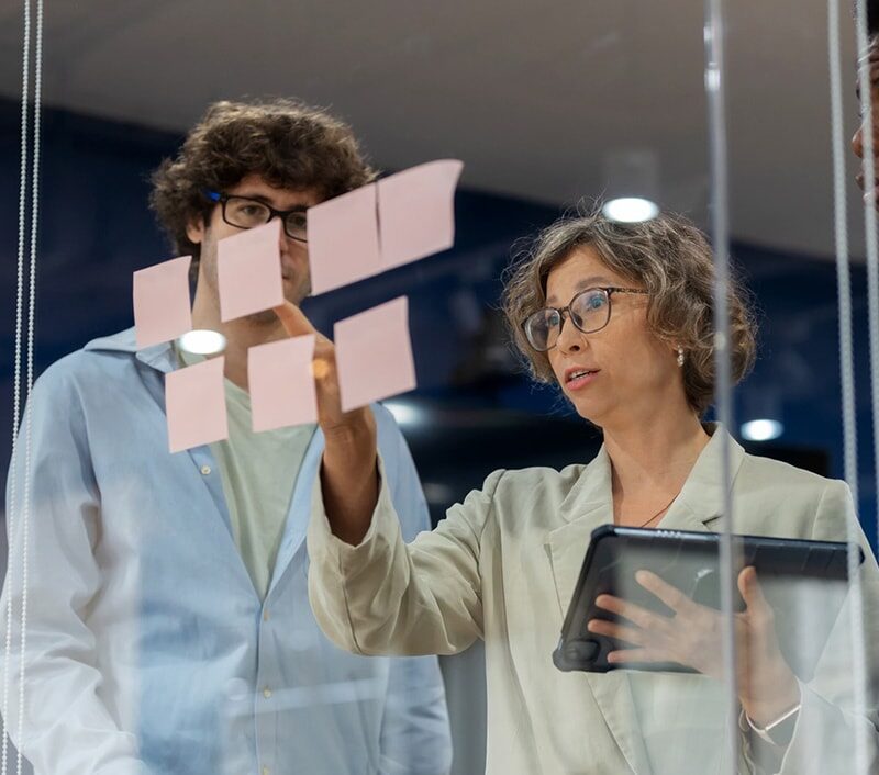 A business development team gathers in a modern office meeting room, brainstorming and discussing their business process improvement strategy. The team, consisting of a diverse group of professionals, uses post-it notes to organize ideas and refine.